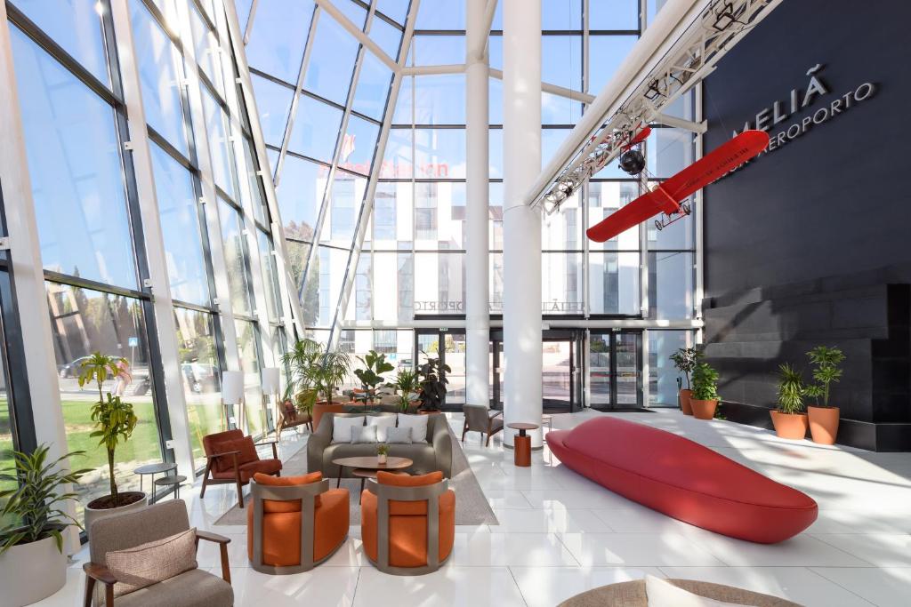 Spacious modern hotel lobby with red decor, large windows, and a hanging airplane model, surrounded by potted plants.