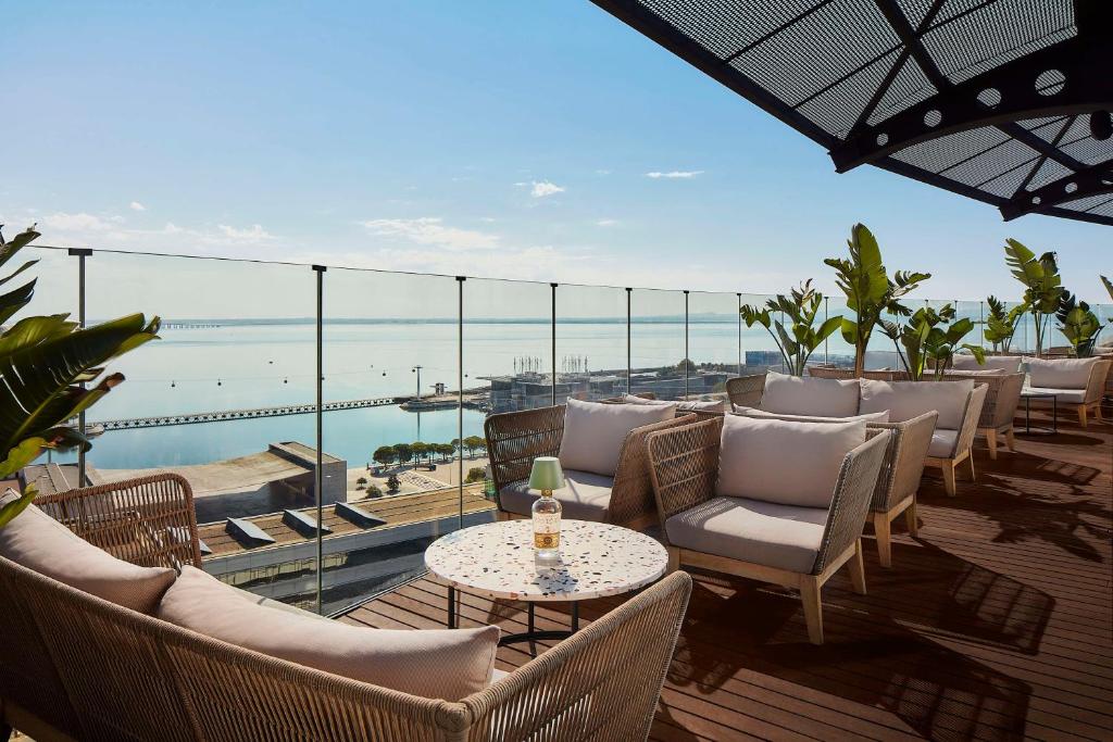 Rooftop lounge with ocean view, wicker chairs, plants, and glass railing under sunny skies. Perfect for relaxation.