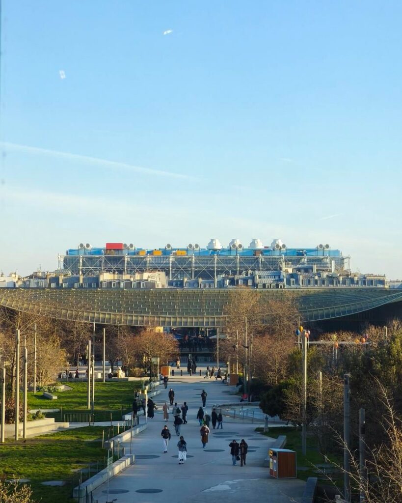 People walking in a park with a modern architectural building in the background under a clear blue sky.