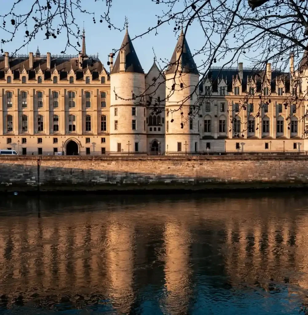 Historic riverside building with towers and reflections, framed by tree branches, in warm sunlight.