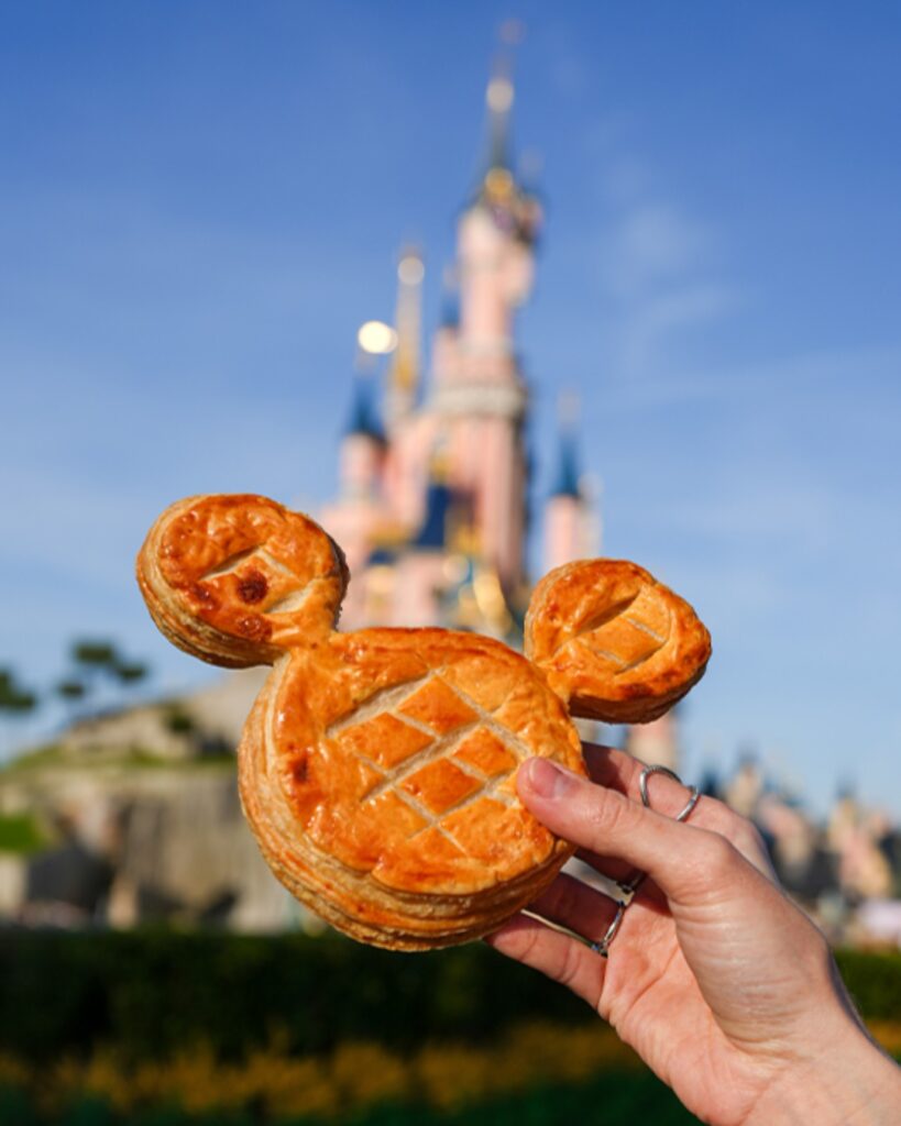 Mickey-shaped pastry held up with castle in the background on a sunny day at an amusement park.