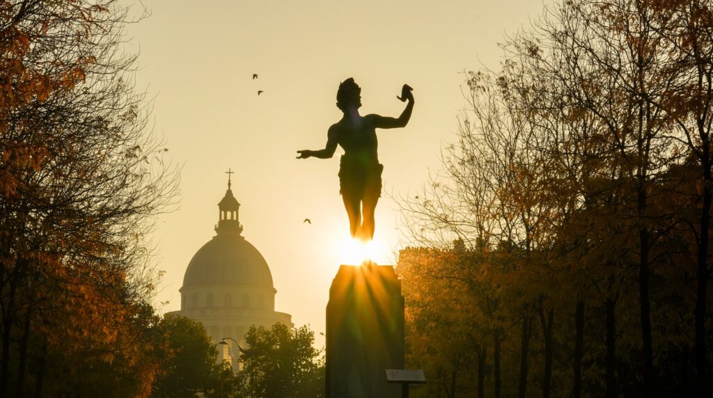 Silhouetted statue at sunrise with a dome and autumn trees in the background.