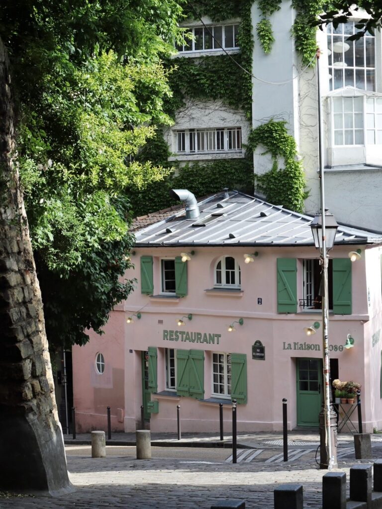 Charming pink French restaurant with green shutters and ivy, nestled in a quaint, tree-lined street.