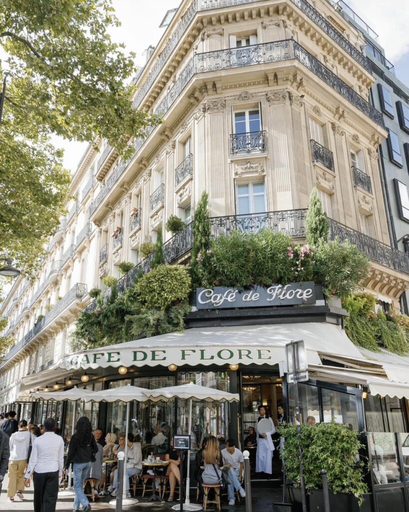 Charming Parisian café with outdoor seating on a bustling street corner, surrounded by greenery and classic architecture.