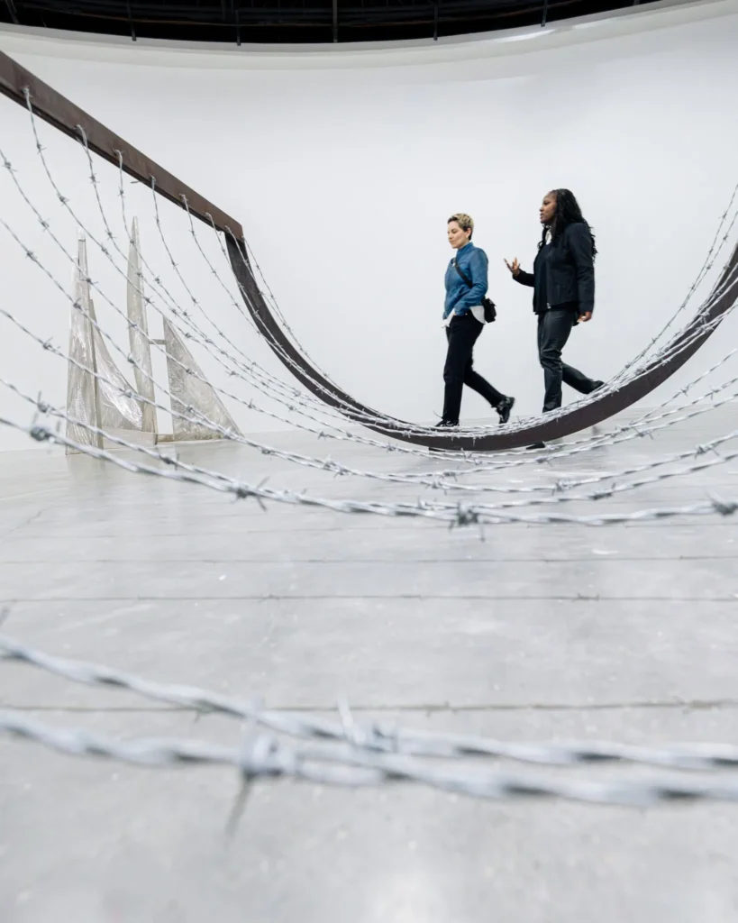Two individuals walking on a curved barbed wire installation in a modern gallery space.
