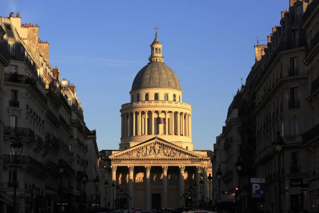Sunlit dome of the Panthéon in Paris between two shadowed streets, under a clear afternoon sky.