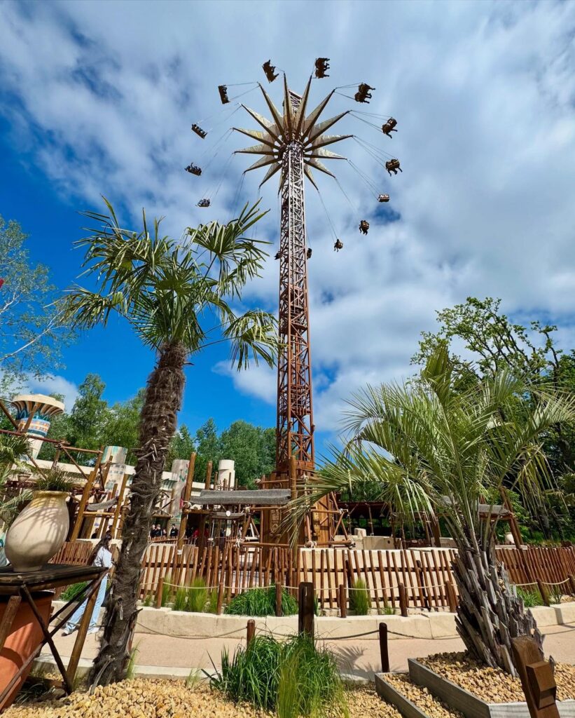 Tall swing ride against blue sky and palm trees in an amusement park setting.