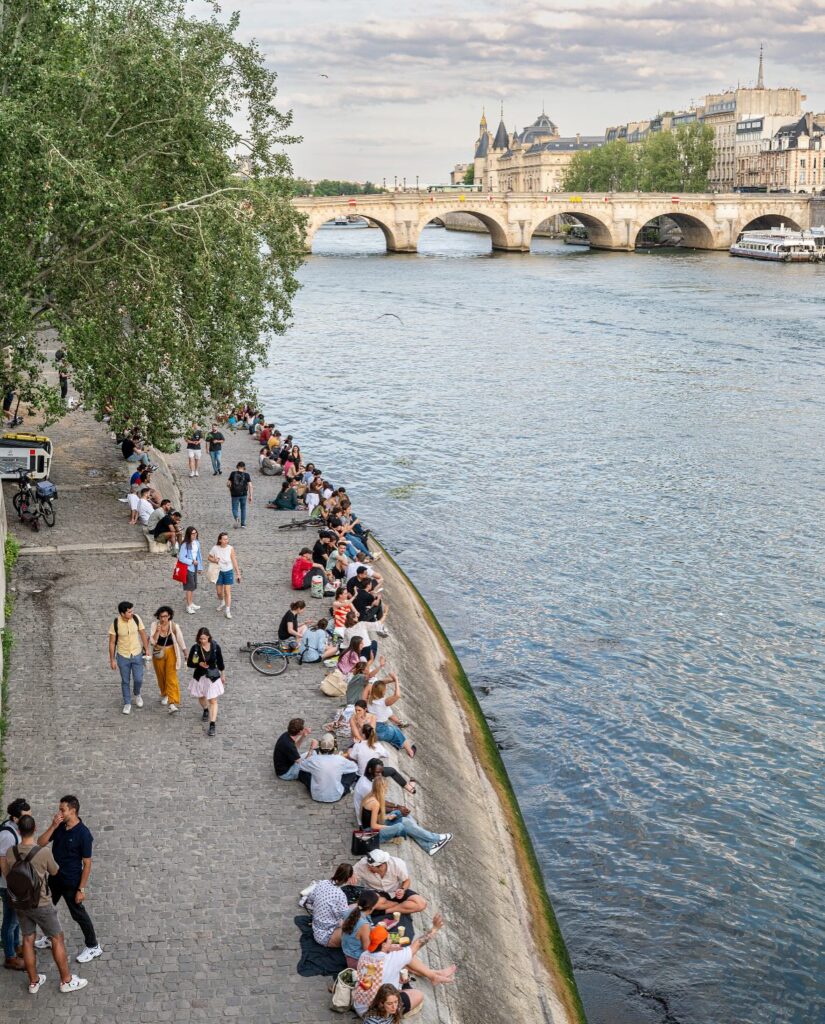 People relaxing by the Seine River in Paris on a sunny day, with a historic bridge in the background.
