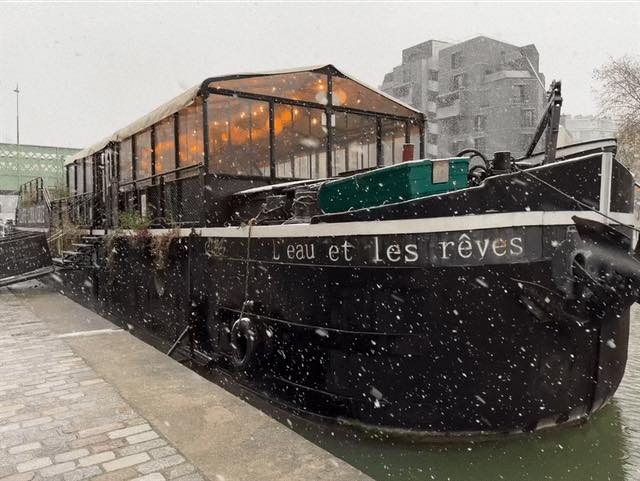 Moored barge in snowfall with a glass-enclosed cabin, urban buildings in the background.