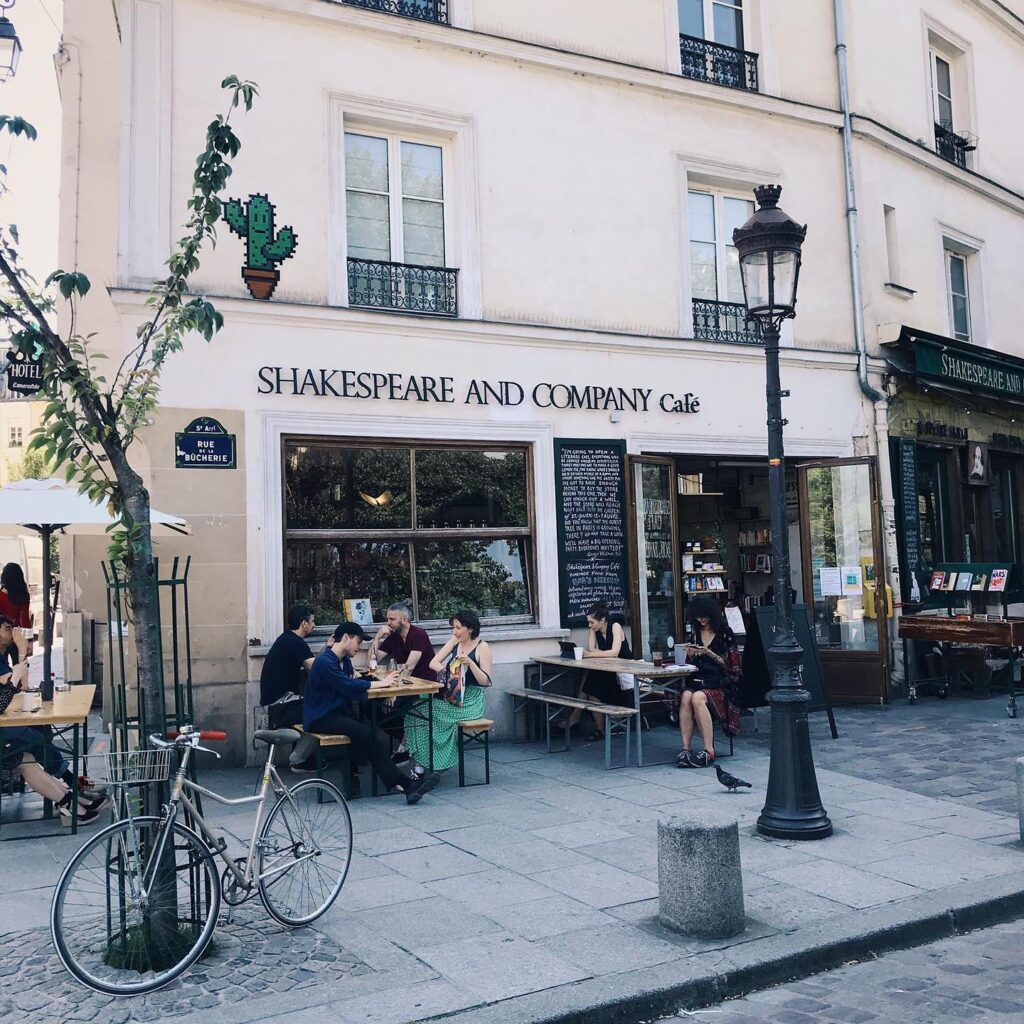 Parisian café scene outside Shakespeare and Company, with people sitting, a bicycle, and charming street ambiance.
