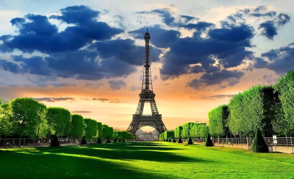 Eiffel Tower at sunset, surrounded by lush green trees and dramatic clouds, Paris icon.