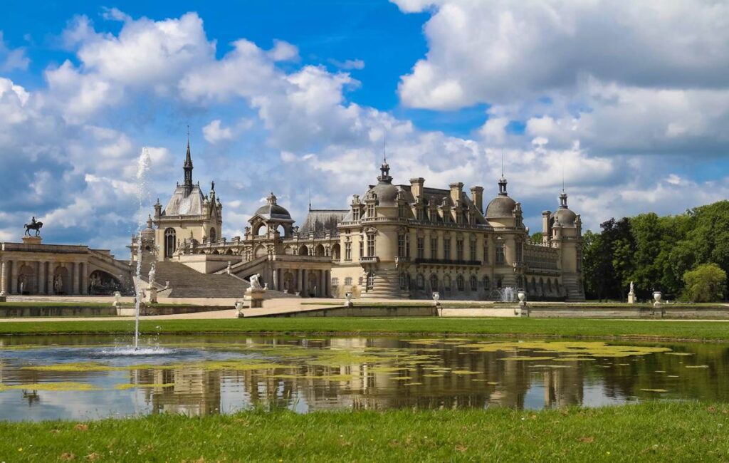 Elegant French chateau reflected in a tranquil pond under a cloudy blue sky, surrounded by lush greenery.