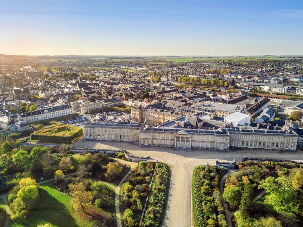 Aerial view of Versailles Palace and gardens, showcasing historic architecture and sprawling landscapes under clear skies.