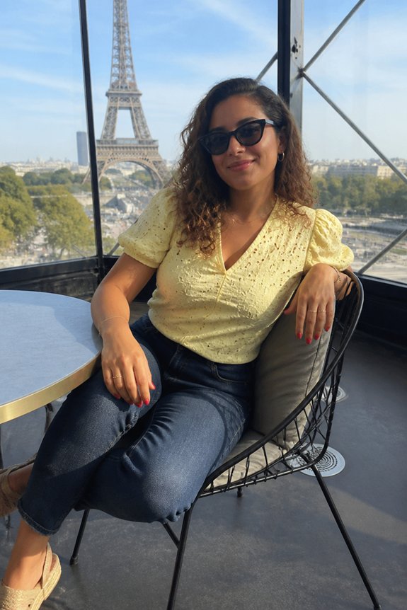 Woman in a yellow top with sunglasses sits at a rooftop Paris cafe with the Eiffel Tower in view.