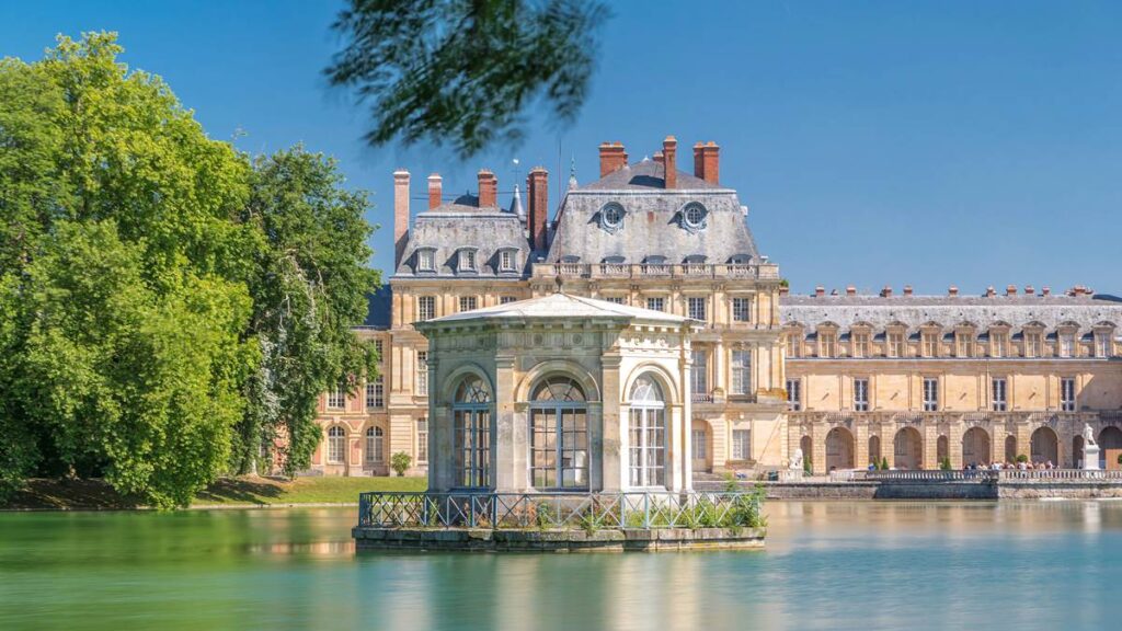 Historic château with elegant pavilion on a serene lake, surrounded by lush greenery under a clear blue sky.