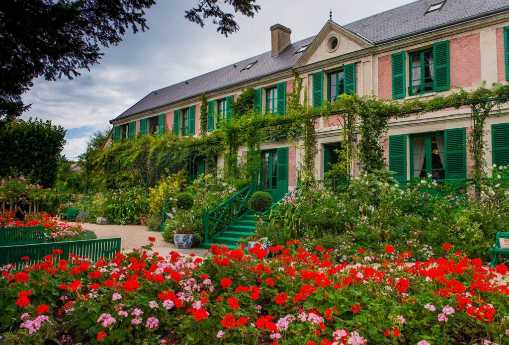 Charming historic house with vibrant garden, red flowers, and green shutters, under a partly cloudy sky.