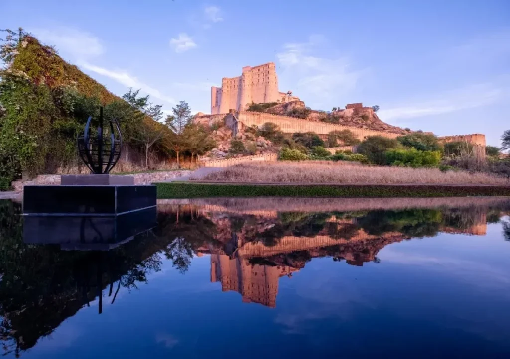 Scenic view of a historic castle reflected in water, surrounded by lush greenery and dramatic sky at sunset.