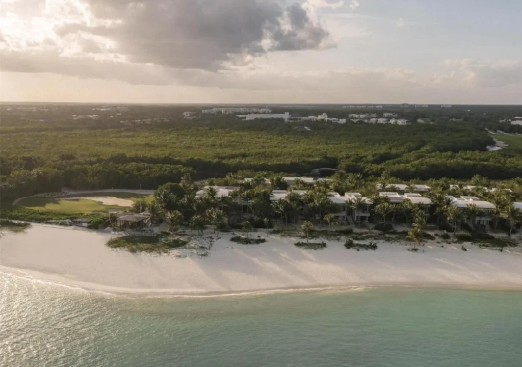 Aerial view of a serene beachfront resort surrounded by lush greenery and calm turquoise waters, under a cloudy sky.
