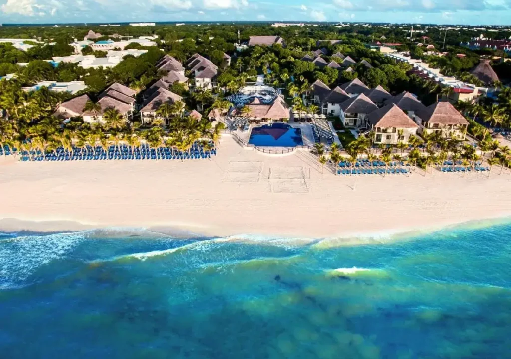 Stunning aerial view of a tropical resort with sandy beach, palm trees, and a pool by the ocean under a clear blue sky.
