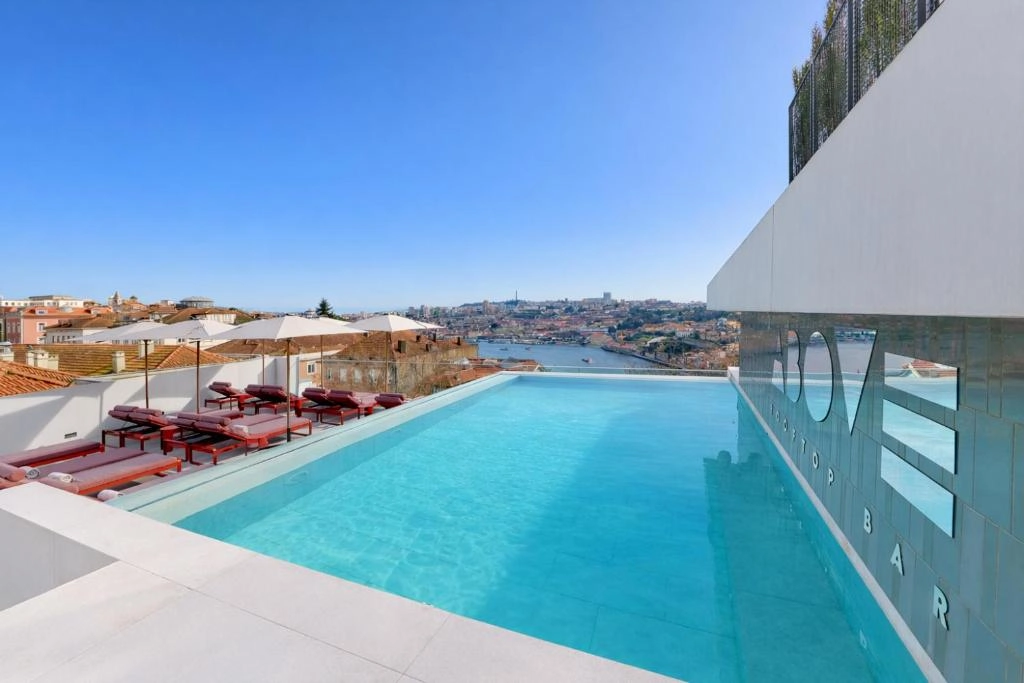 Rooftop pool with stunning city views, lounge chairs, and umbrellas at a stylish bar under a clear blue sky.