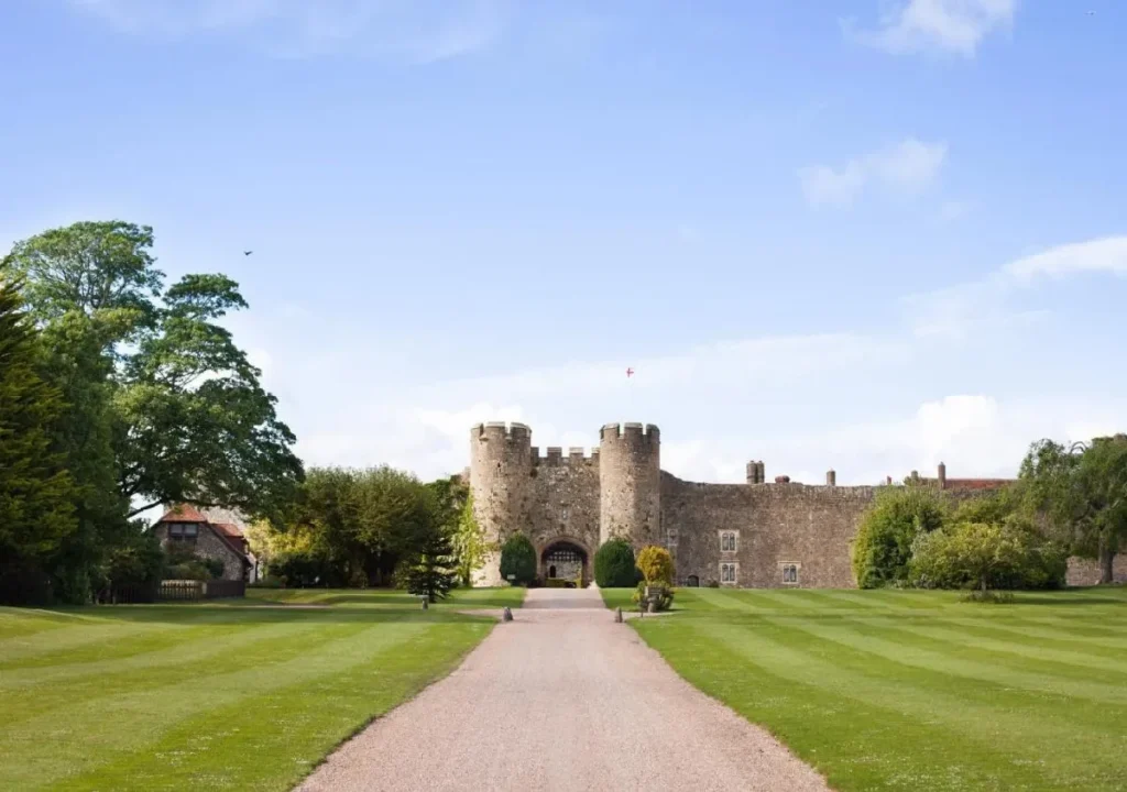 Scenic view of a historic castle entrance with manicured lawns and trees lining the pathway under a clear blue sky.