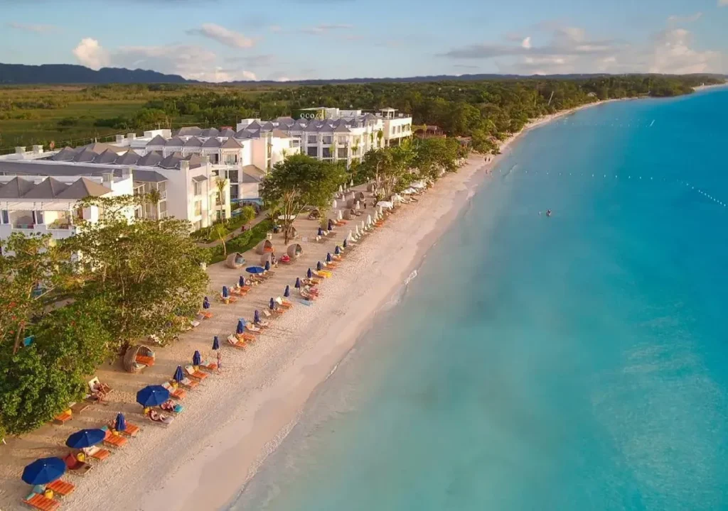 Serene beach view with lounge chairs, umbrellas, and a resort along turquoise waters and lush greenery. Perfect for relaxation.
