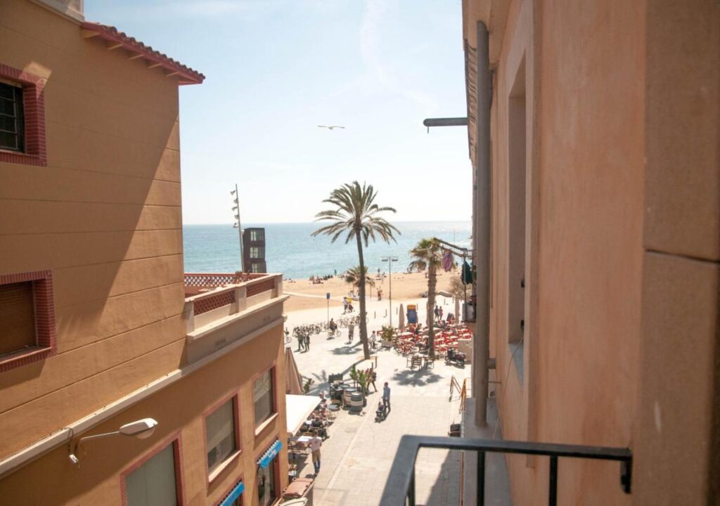 View from a balcony overlooking a sunny beach, palm trees, and people enjoying seaside activities.