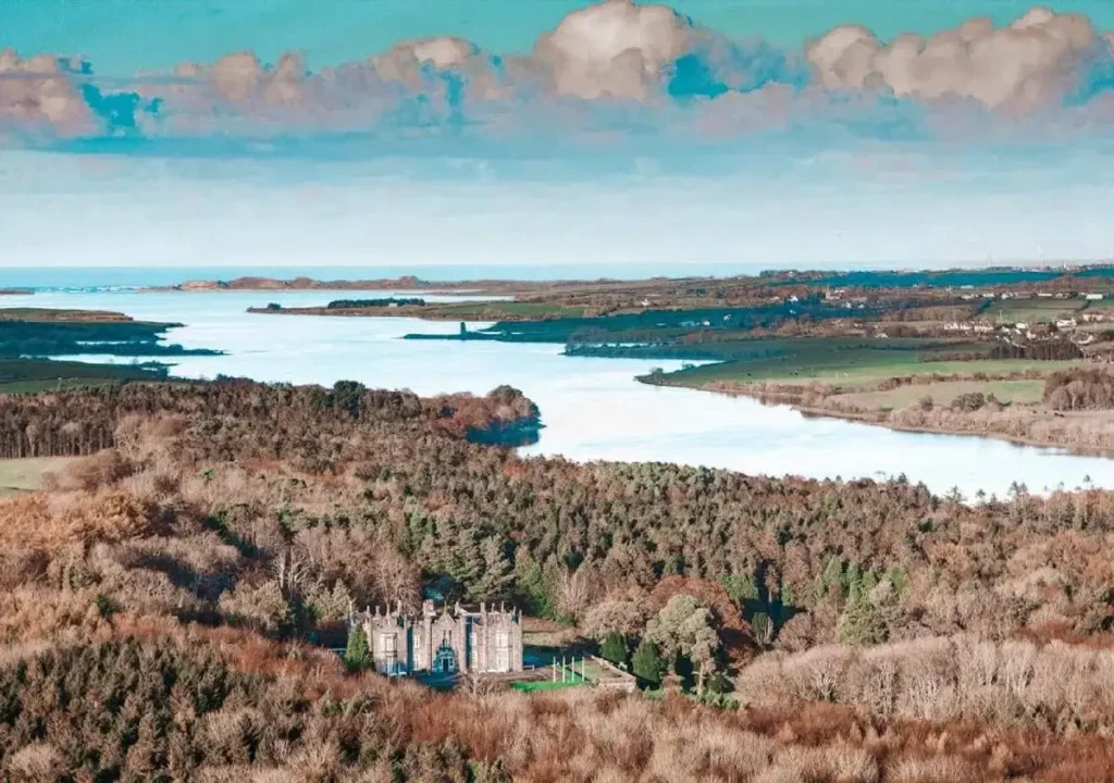 Scenic aerial view of a majestic castle surrounded by lush greenery and a winding river under a clear blue sky.