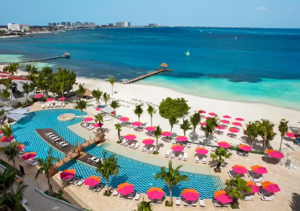 Vibrant beachside resort with a pool, lounge chairs, pink umbrellas, and turquoise water under a clear blue sky.