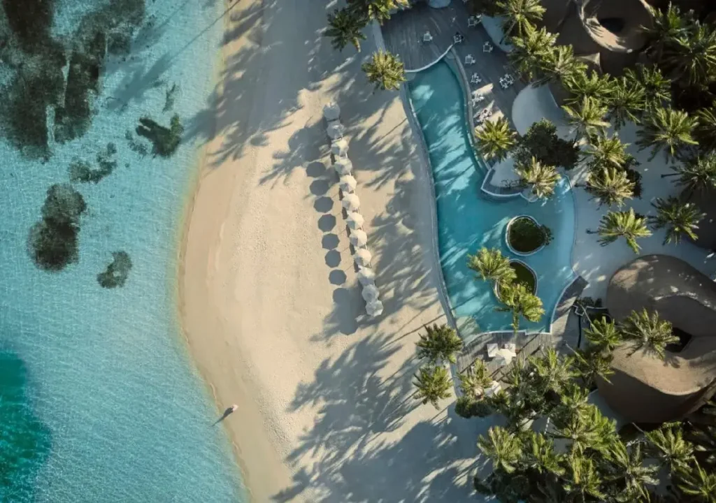 Aerial view of a serene beach with a swimming pool, palm trees, and lounge chairs under sunny skies. Perfect vacation spot.