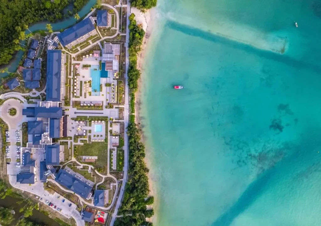 Aerial view of a luxury resort with a landscaped pool and beach area next to turquoise water and lush greenery.
