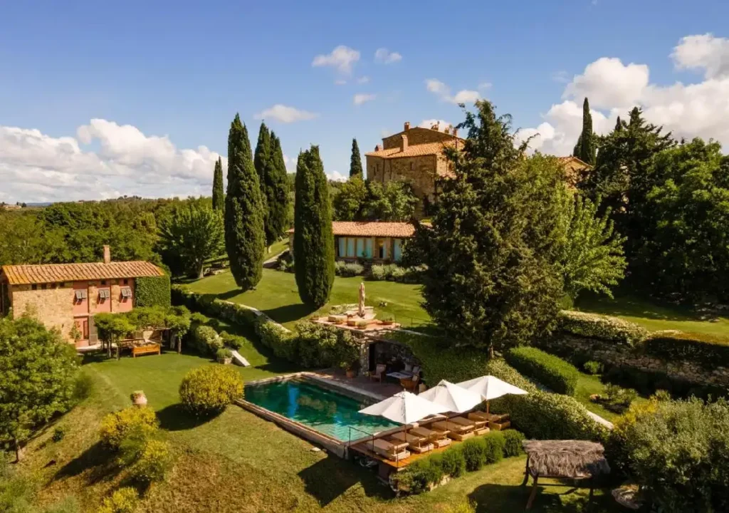 Alt text: Scenic Tuscan villa featuring a lush garden, swimming pool, and cypress trees under a clear blue sky.