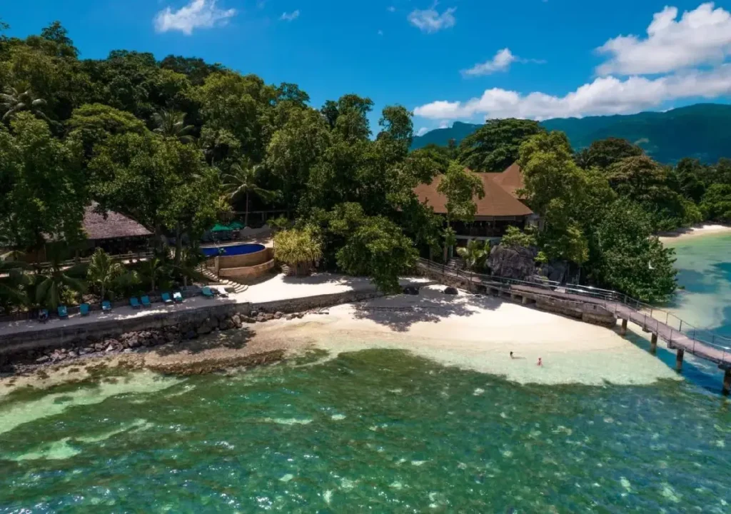 Aerial view of a tropical beach with clear water, lush greenery, and a villa surrounded by palm trees. Relaxing paradise getaway.