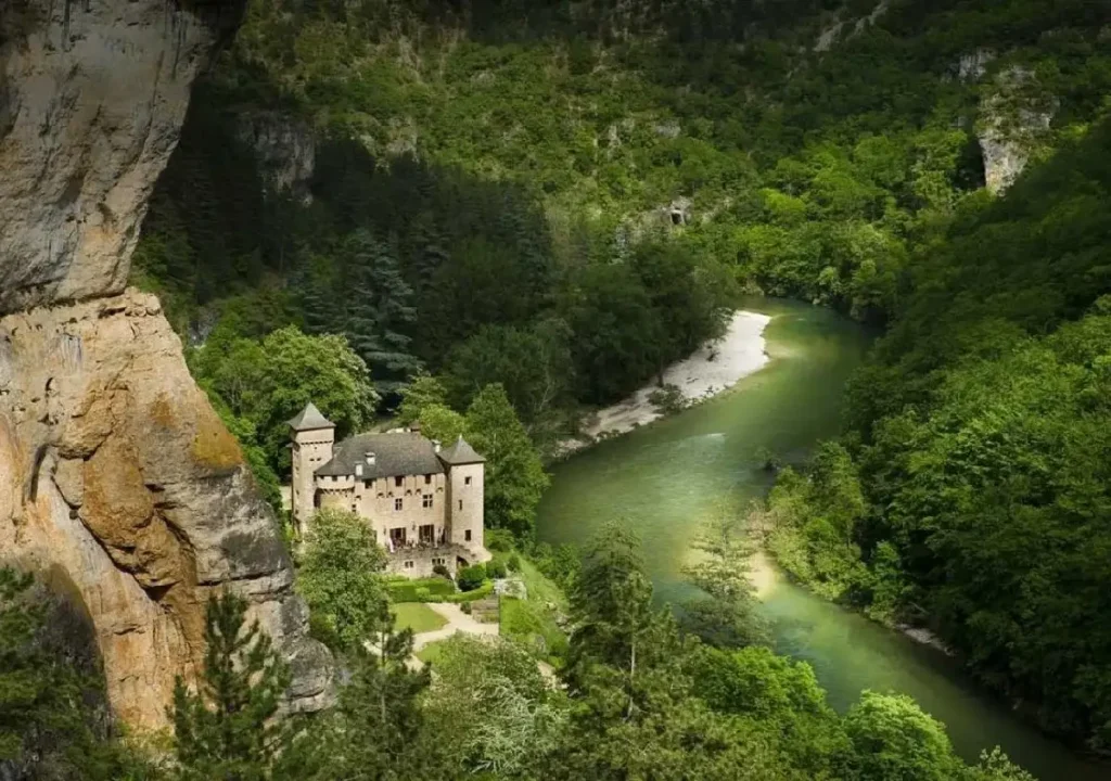 Scenic view of a castle nestled in lush greenery by a winding river, surrounded by rocky cliffs and dense forests.