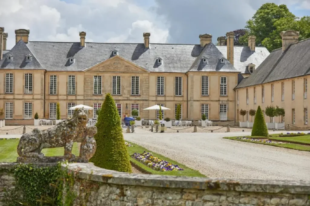 Elegant historic chateau with manicured gardens, featuring a lion statue and outdoor seating area under umbrellas.