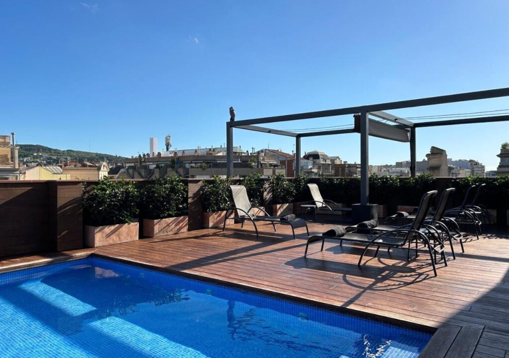 Rooftop pool with sun loungers and greenery, featuring a clear blue sky above the city skyline. Perfect for relaxation.