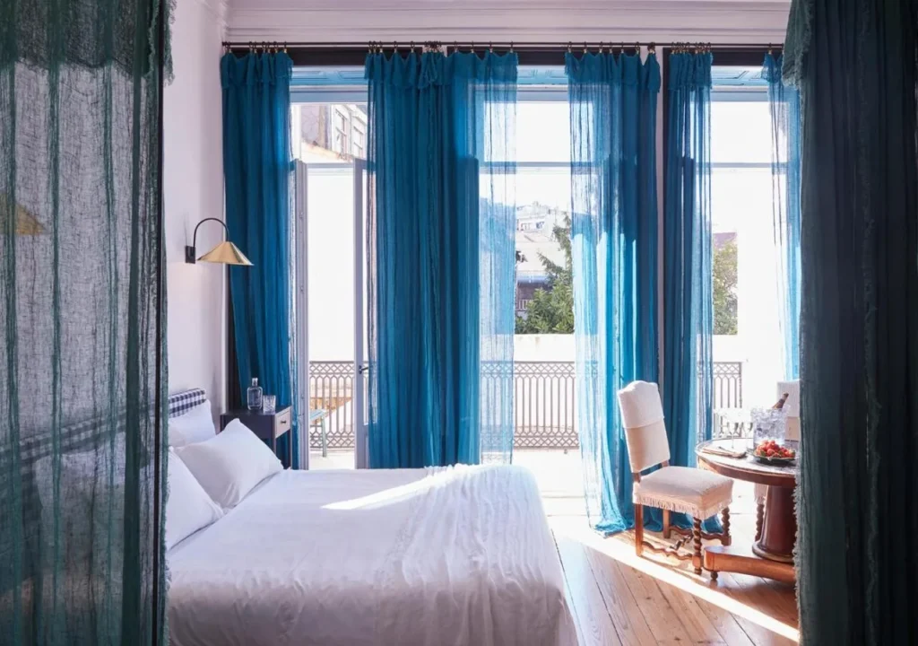 Bright and airy bedroom featuring large windows with turquoise curtains, a cozy bed, and a charming sitting area.