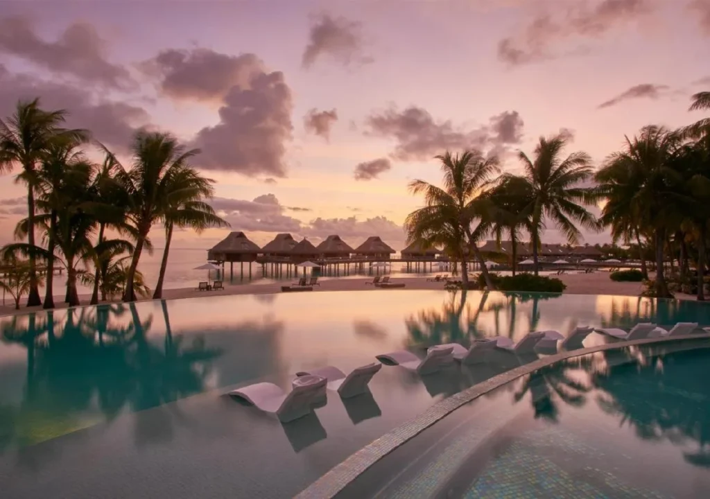 Serene oceanfront resort pool at sunset, surrounded by palm trees and overwater bungalows reflecting golden light.