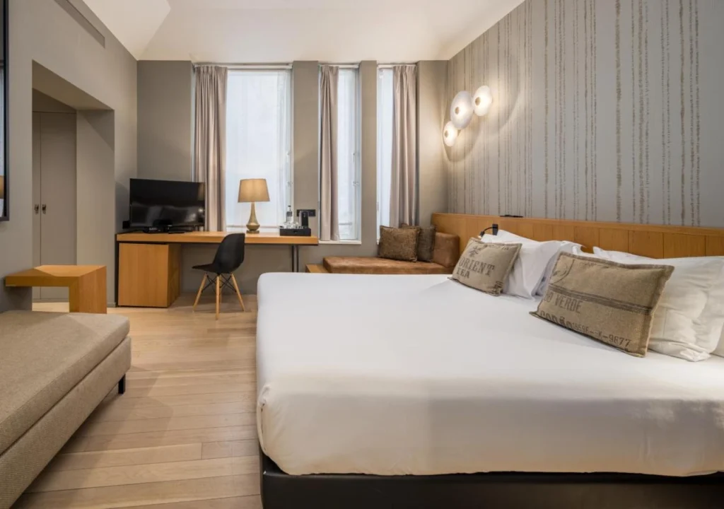 Modern hotel room featuring a spacious bed, desk area, and cozy seating with natural light from large windows.