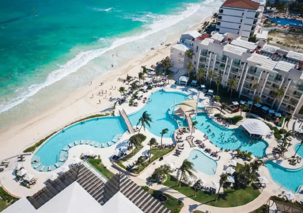 Aerial view of a beach resort with pools, palm trees, and beachgoers enjoying the sun by the turquoise ocean.