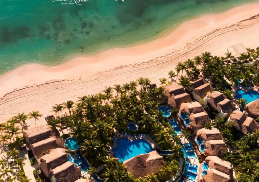 Aerial view of a tropical resort with sandy beach, palm trees, and sparkling pools next to turquoise waters.