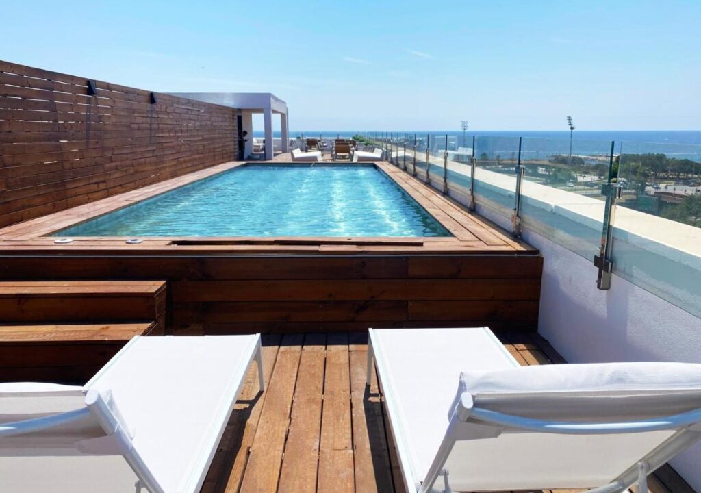 Rooftop pool with wooden deck and lounge chairs, overlooking the ocean on a sunny day. Perfect for relaxation and adventure.