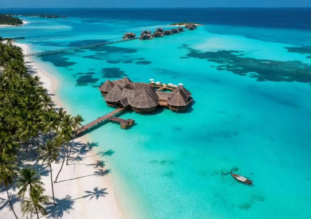 Aerial view of a tropical resort on turquoise waters with palm trees and a wooden walkway leading to overwater bungalows.