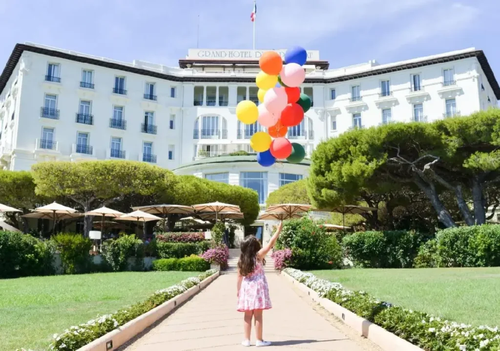 A child in a floral dress holds colorful balloons outside the Grand Hotel, surrounded by lush gardens and umbrellas.