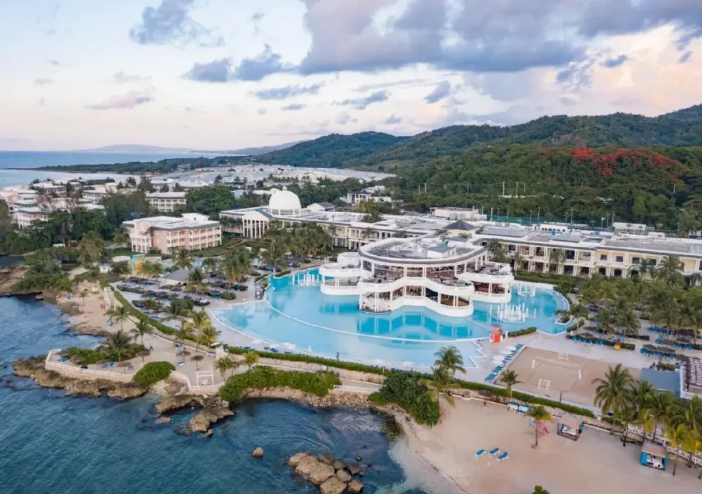 Aerial view of a luxurious resort with a large pool, beach, and palm trees against a scenic hillside backdrop.