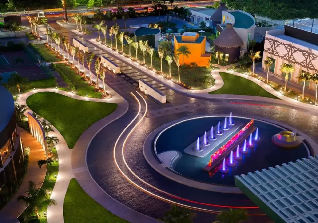 Night view of a landscaped resort entrance featuring colorful fountains, palm trees, and illuminated pathways.