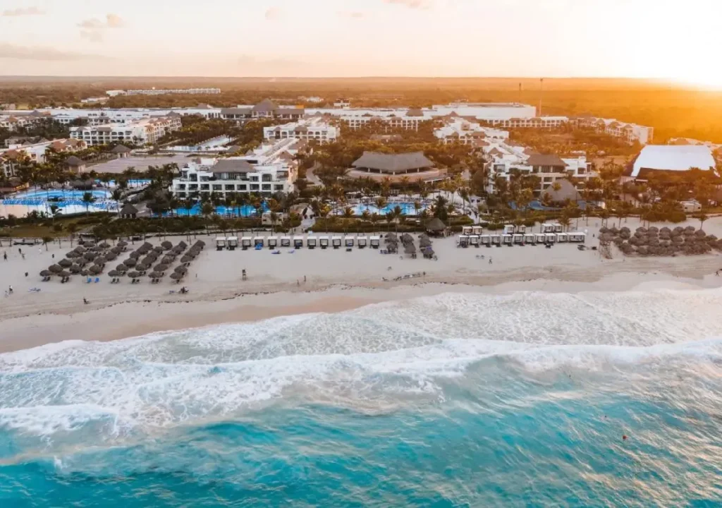 Aerial view of a beachfront resort in Cancun at sunset, featuring pools, beach cabanas, and palm trees.