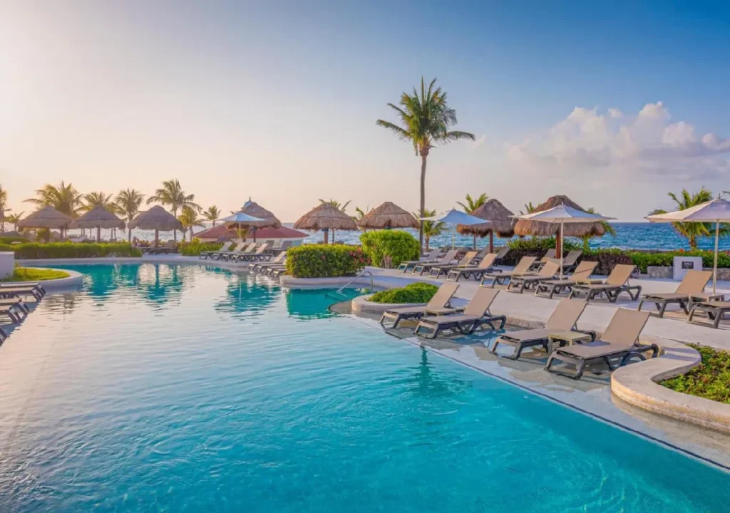 Serene resort pool with loungers and thatched umbrellas, framed by palm trees and ocean views at sunset.