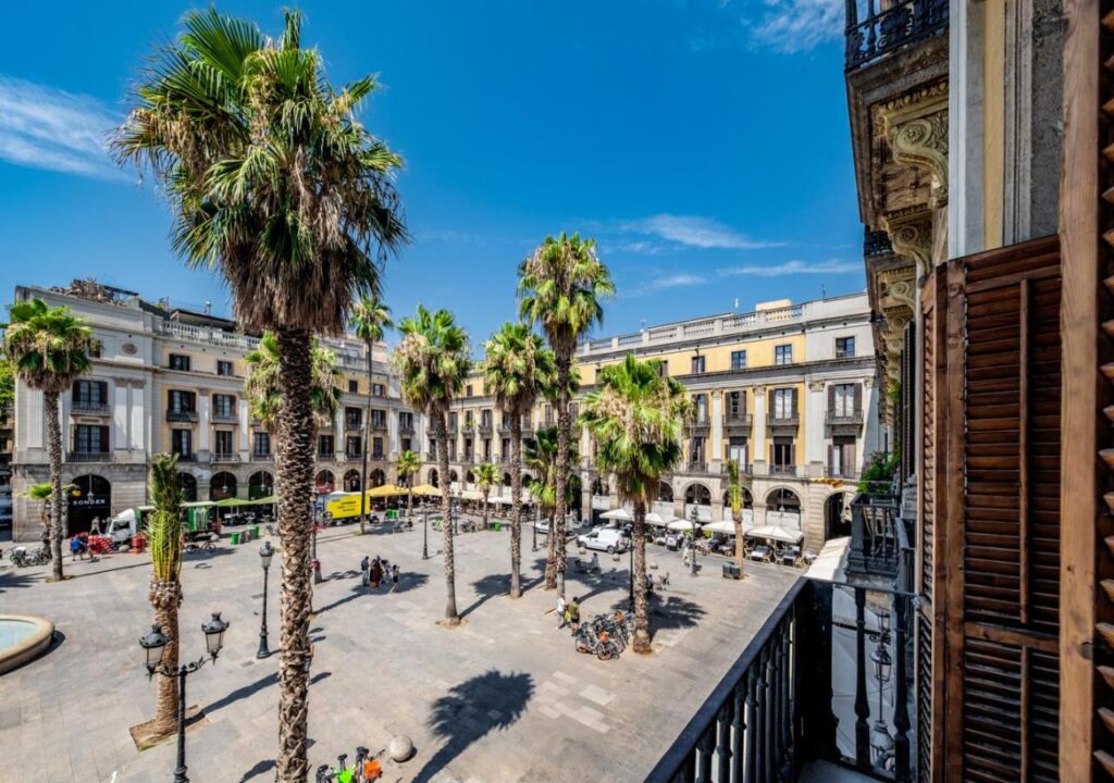 View from a balcony overlooking a vibrant plaza with palm trees, cafes, and clear blue skies in Barcelona.