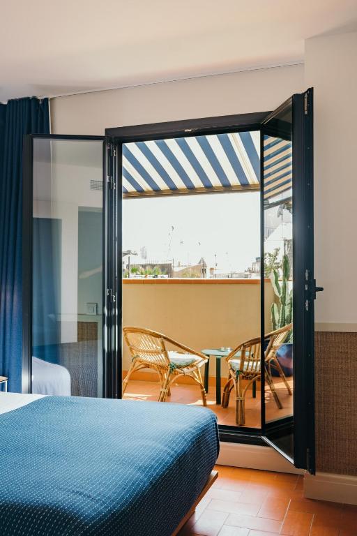 Cozy hotel room with a blue bedspread, glass doors opening to a balcony with chairs and a table, and striped awning.